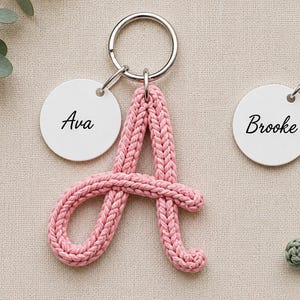 May include: A keychain featuring a pink knitted letter A attached to a silver metal ring and a round white disc with the name Ava printed in black script. A second round white disc with the name Brooke printed in black script is also visible.