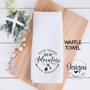 May include: White waffle towel with the text "NEW HOME new Adventure NEW MEMORIES" in a circular design. The towel also has a key and house graphic. The words "WAFFLE TOWEL" are printed vertically on the right side of the towel.
