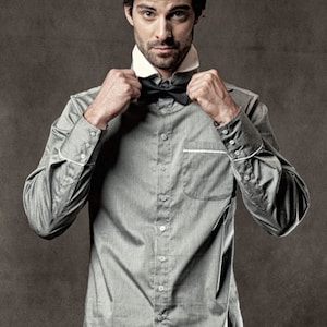 May include: A man wearing a gray button-down shirt with a black bow tie. He is adjusting the bow tie with both hands.