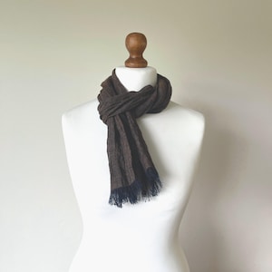 May include: A dark brown scarf with a textured weave and dark blue fringe at the ends. The scarf is wrapped around a white mannequin neck, displaying a simple knot style with the two ends hanging down in front of the chest area.