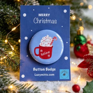 Peut inclure: Un badge rond sur une carte de présentation bleue avec le texte Merry Christmas, Button Badge et Lazymitts.com. Le badge présente une tasse rouge avec le mot Believe, remplie de crème fouettée et un sucre d'orge sur un fond bleu clair.