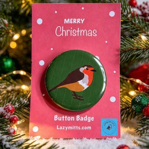 May include: A circular button badge featuring a robin illustration on a dark green background, mounted on a red card printed with white snowflakes and the text Merry Christmas, Button Badge, and Lazymitts.com with a small blue logo.