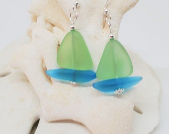 May include: A pair of sailboat-shaped earrings. The earrings feature a light green sail and a blue hull, connected by silver-colored hooks. The earrings are displayed on a white, textured surface.