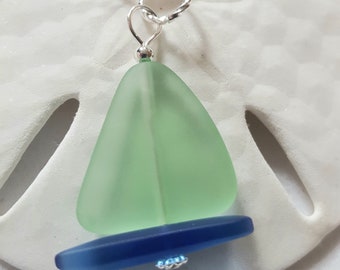 May include: A sea glass sailboat pendant necklace with a light green sail and a dark blue hull. The pendant is attached to a silver chain.