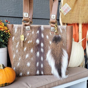 May include: A brown leather tote bag with a spotted fawn skin design. The bag has gold star accents and a brown fur keychain. The bag is sitting on a brown bench with a pumpkin and flowers in the foreground.