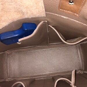 May include: The inside of a brown leather bag with multiple pockets. A blue handgun is visible in one of the pockets.