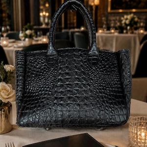 May include: A black handbag with a prominent crocodile skin texture, featuring two sturdy top handles and a structured rectangular body. The surface displays a detailed, raised scale pattern across the entire exterior, providing a distinct tactile appearance.