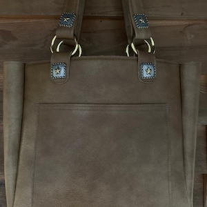 May include: A brown leather tote bag with a large pocket on the front. The bag has two top handles with gold rings and star-shaped accents.