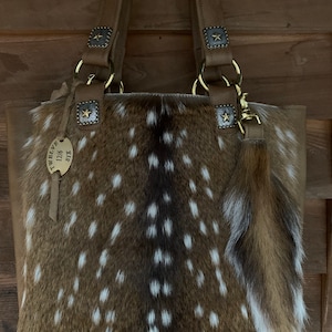 May include: A brown leather tote bag with a spotted fawn fur body and gold hardware. The bag has a small, brown leather tassel with a gold star charm. The bag has a tag that reads "Twelve 0116 SX".