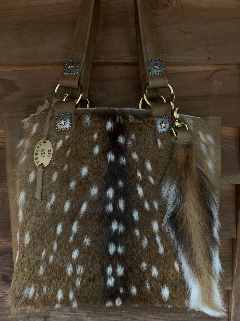 May include: A brown leather tote bag with a spotted fawn fur body and gold hardware. The bag has a small, brown leather tassel with a gold star charm. The bag has a tag that reads "Twelve 0116 SX".
