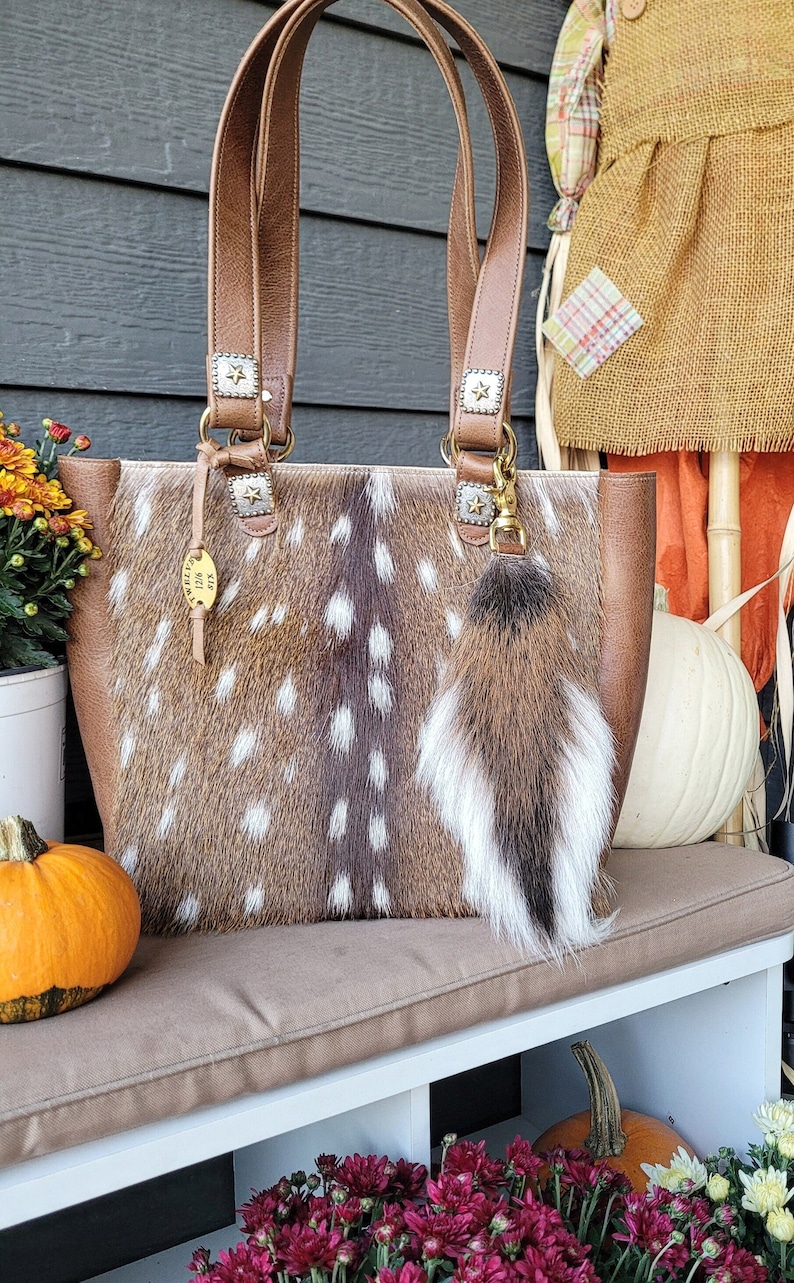 May include: A brown leather tote bag with a spotted fawn skin design. The bag has gold star accents and a brown fur keychain. The bag is sitting on a brown bench with a pumpkin and flowers in the foreground.
