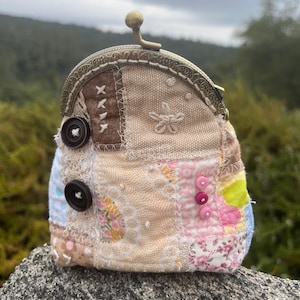 May include: A patchwork coin purse with a brass clasp and two large buttons. The purse features a variety of fabrics in beige, brown, pink, and blue, with embroidered details and decorative stitching. The purse is sitting on a rock.