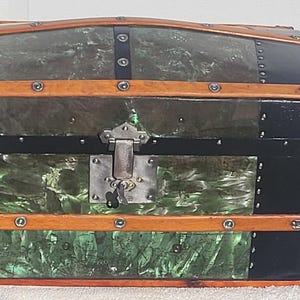 May include: An antique trunk with a domed top, featuring black, green, and wood-toned panels. The trunk has metal hardware and a lock. A can of Sunkist Zero Sugar Orange Soda is next to the trunk.