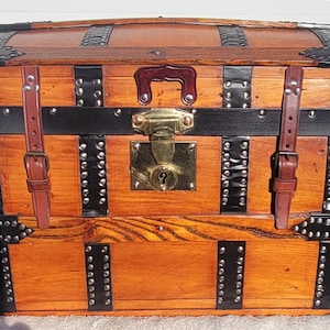 May include: A rectangular wooden storage chest with a domed lid, featuring natural wood grain, black metal corner reinforcements, and vertical black metal bands secured with silver-toned rivets. It includes two brown leather straps and a brass latch.