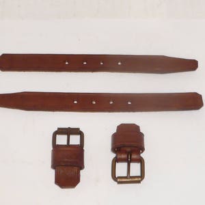 May include: Two brown leather straps with brass buckles. The straps are approximately 1 inch wide and have four evenly spaced holes.