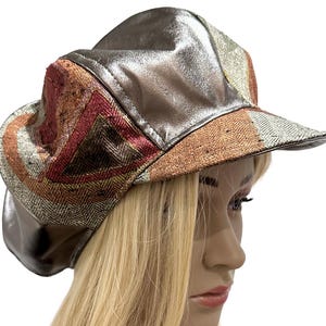 May include: A multi-textured newsboy cap featuring panels of metallic silver fabric, brown herringbone tweed, and geometric patterns in shades of rust, orange, and cream. The hat has a structured brim and a rounded, paneled crown construction.