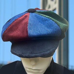 May include: A multi-colored newsboy cap featuring a patchwork design with panels in shades of red, blue, and green. The hat is constructed from a textured corduroy-like fabric and includes a solid dark gray brim and a small matching fabric button.