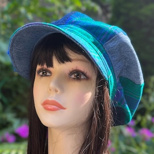 May include: A newsboy cap featuring a patchwork design of blue and green plaid fabric combined with solid grey denim panels. The hat has a structured brim and a rounded crown, showcasing a mix of textures and colors in a classic multi-panel construction.