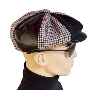 May include: An eight-panel newsboy cap featuring alternating sections of black leather and a beige and black houndstooth patterned fabric. The cap has a structured black leather brim and a small fabric-covered button at the center of the crown.