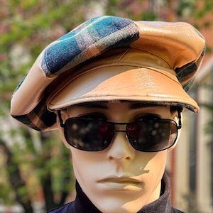 May include: A newsboy cap featuring a combination of tan leather and a blue, green, and black plaid fabric pattern. The cap has a structured tan leather brim and a rounded crown. The cap is worn by a mannequin wearing black-framed sunglasses.