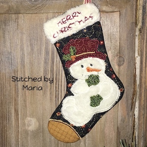 May include: A black and white Christmas stocking with a snowman design. The stocking has a white fur cuff with the text "Merry Christmas" embroidered in red. The snowman is wearing a red hat with green trim and a green scarf. The stocking is hanging on a wooden surface.