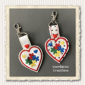 May include: Two white heart-shaped keychains with red stitching. Each keychain features a red button and a colorful puzzle piece design. The keychains are attached to silver key rings. The text "NonnieJoo Creations" is visible at the bottom of the image.