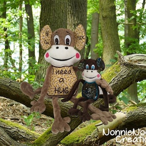 May include: Two plush monkey toys sitting on a tree branch. The larger monkey is brown with a yellow and black patterned shirt that says "I need a hug". The smaller monkey is brown with a blue and black patterned shirt.
