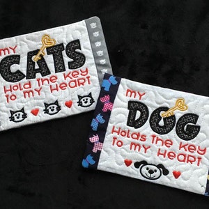 May include: Two white fabric coasters with embroidered designs. The first coaster has a black cat design with the text "MY CATS Hold the key to MY HEART". The second coaster has a black dog design with the text "MY DOG Holds the key to MY HEART".