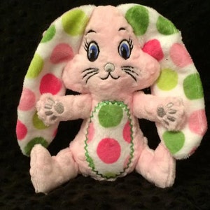Puede incluir: Un conejito de peluche rosa con lunares verdes y rosas. El conejito tiene ojos azules y una nariz blanca.