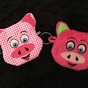 May include: Two pink pig-shaped plush keychains. One keychain is pink gingham with a green zipper pull. The other keychain is a fuzzy pink pig with green ears.