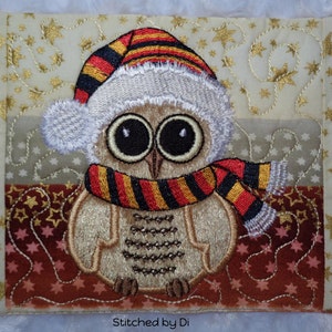 May include: A brown and white owl wearing a red and black striped hat and scarf. The owl is embroidered on a beige fabric with gold stars and a red fabric with pink stars. The text "Stitched by Di" is at the bottom of the image.