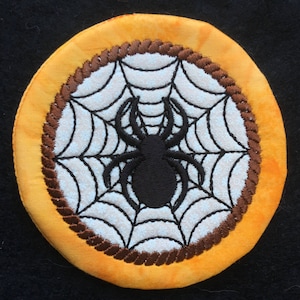Può includere: Sottobicchiere rotondo a tema Halloween con un ragno nero al centro di un disegno a ragnatela bianca. La ragnatela è posta su uno sfondo chiaro, circondata da un bordo marrone a forma di corda e da un bordo in tessuto giallo-arancio. Per Halloween.