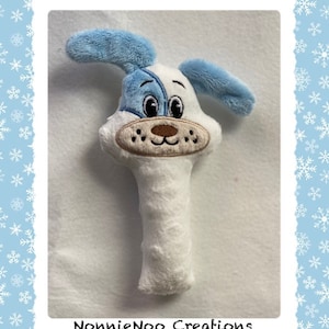 Op de afbeelding: Een witte en blauwe pluche hondenrammelaar met geborduurde ogen en een bruine neus. De rammelaar is ontworpen voor baby's en peuters.