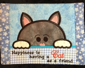 May include: A blue and white checkered fabric coaster with a gray cat peeking over the top. The cat has pink ears and a pink nose. The text "Happiness is having a Cat as a friend" is embroidered on the coaster.