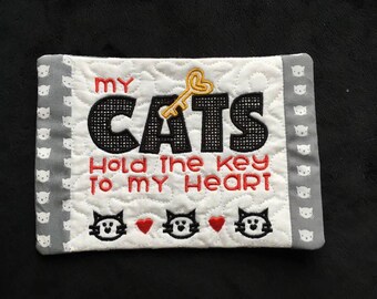 May include: White quilted coaster with gray border featuring black embroidered text "My Cats Hold The Key To My Heart" with a gold key and three black cat faces with red hearts.