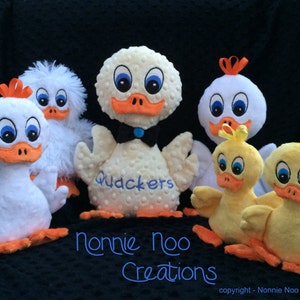 May include: Five plush toy ducks in various colors and sizes. The ducks are all white with orange beaks and blue eyes. One duck is larger than the others and has the word "Quackers" embroidered on its chest. The ducks are arranged in a semi-circle on a black background.