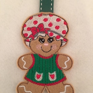 May include: A gingerbread person ornament with a red and white polka dot hat, a green dress with red hearts, and a white bow. The ornament is made of felt and has a green ribbon for hanging.