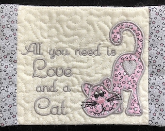 May include: A white quilted coaster with pink and gray stars on the border. The coaster has a gray embroidered message that reads "All you need is Love and a Cat". A pink cat with a heart on its back is embroidered on the coaster.