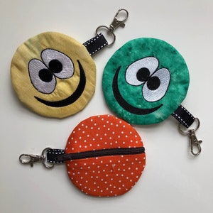 Könnte beinhalten: Drei kleine, runde Reißverschlusstaschen mit Schlüsselanhängern. Die Taschen sind aus Stoff und haben verschiedene Designs: ein gelbes Smiley-Gesicht, ein grünes Smiley-Gesicht und ein rotes Punkte-Muster.