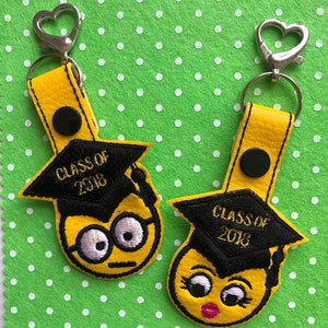 May include: Two yellow graduation keychains with black graduation caps and the text "Class of 2018". One keychain features a yellow emoji with glasses and a neutral expression. The other keychain features a yellow emoji with eyelashes, a red lipstick, and a slightly surprised expression.