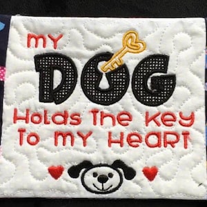 May include: A white quilted coaster with a black and yellow embroidered keyhole and the text "My Dog Holds the Key to My Heart". The coaster is surrounded by a blue fabric border with a repeating pattern of small, colorful dogs.
