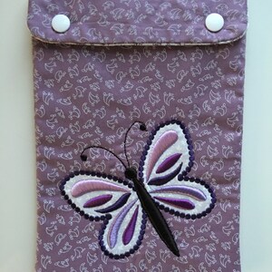 May include: A lavender fabric case with a white floral pattern and a large embroidered butterfly. The butterfly has white, lavender, and purple details. The case has a flap closure with two white buttons.