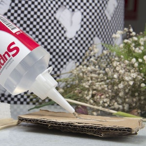 May include: A clear glue bottle with a red and white label is dispensing glue onto a piece of corrugated cardboard. The glue is being applied in a thin line. The background includes a black and white checkered pattern and dried flowers.