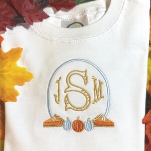 Könnte beinhalten: Weißes T-Shirt mit einem gestickten Design mit den Buchstaben "JSM" in Gold, innerhalb eines hellblauen Ovals. Das Design umfasst zwei Kuchenstücke und drei Kürbisse in Orange- und Blautönen. Das T-Shirt ist von Herbstblättern umgeben.
