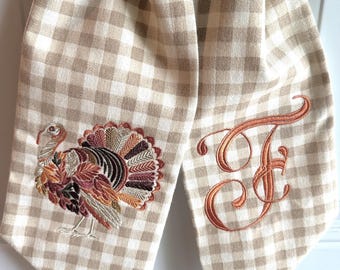 May include: A beige and white checkered kitchen towel with a detailed embroidered turkey and the letter "F" in a copper color. The towel is folded in half, creating a decorative hanging loop.