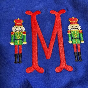 Puede incluir: Camiseta azul real con una gran letra "M" roja bordada flanqueada por dos figuras de cascanueces bordadas. Los cascanueces son verdes, rojos, amarillos y negros. La camiseta está hecha de una tela suave y texturizada.