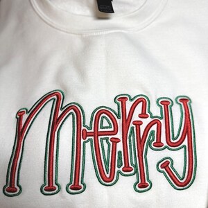 May include: A white sweatshirt with the word "Merry" embroidered in red with green outlines. The festive design is perfect for the holiday season. The sweatshirt is made of a soft material.