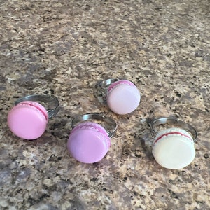 May include: Four silver rings with different colored macarons attached. The macarons are pink, purple, white, and light pink.