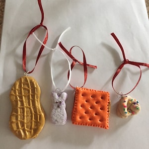 May include: Four handmade Christmas ornaments shaped like a peanut, a bunny, a cracker, and a sprinkle-covered pastry. Each ornament is attached to a red ribbon.