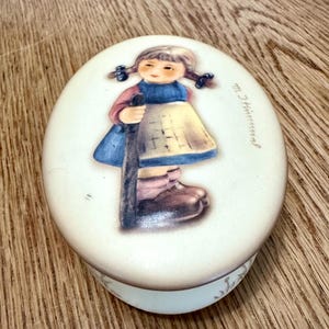 May include: Oval ceramic trinket box with a painted illustration of a young girl in a blue dress, yellow apron, and brown boots, holding a staff. The box has a cream-colored lid and base, with the name "M. Hummel" written on the side.